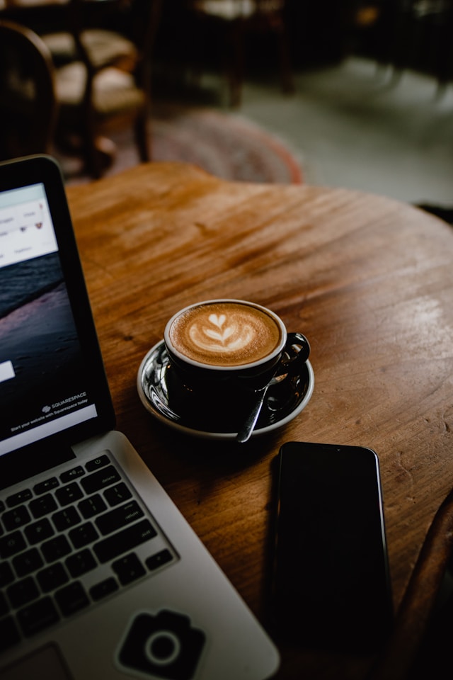 A cup of coffee in a black ceramic cup beside a laptop on a café table