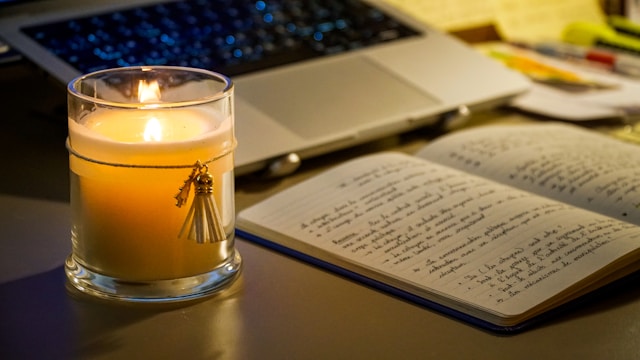 A lit candle beside an open notebook and a laptop
